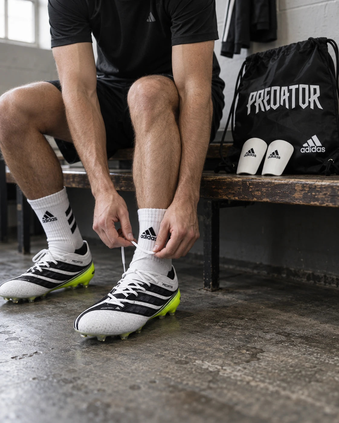 Joueur laçant ses crampons Adidas Predator, avec un sac à crampons et des protège-tibias sur le banc du vestiaire.